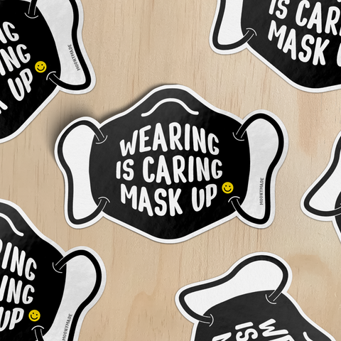 Stickers of a black face mask illustration with 'Wearing is Caring Mask Up' text on a wooden surface.