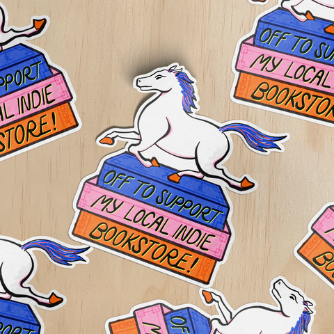 Stickers with horses galloping on a stack of books. Text on the spines of the books reads, 'Off to support my local indie bookstore!' on a wood surface.